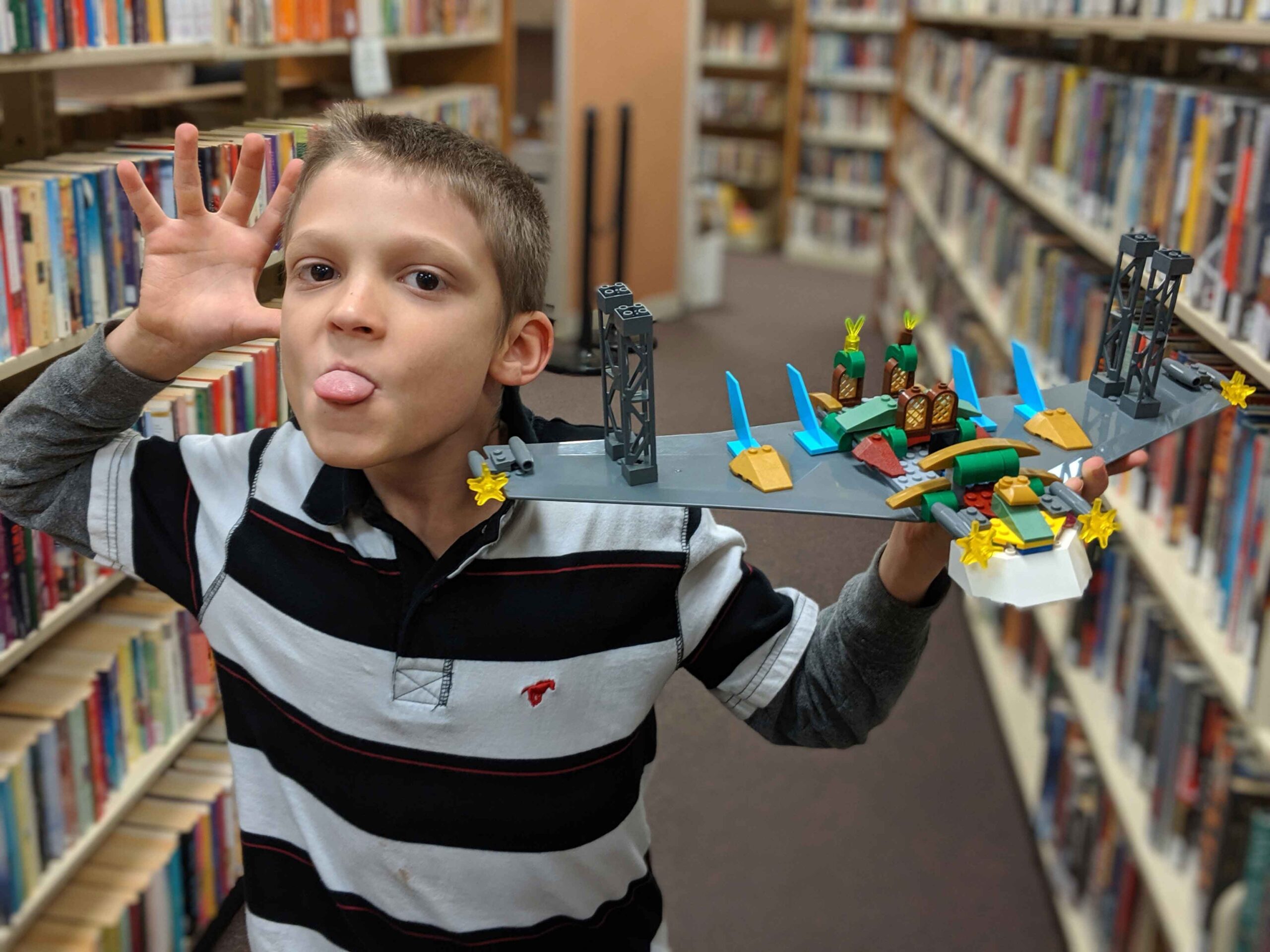 Young boy in a striped shirt holds up his LEGO creation while making a funny face. In the background are shelves of books in a library.